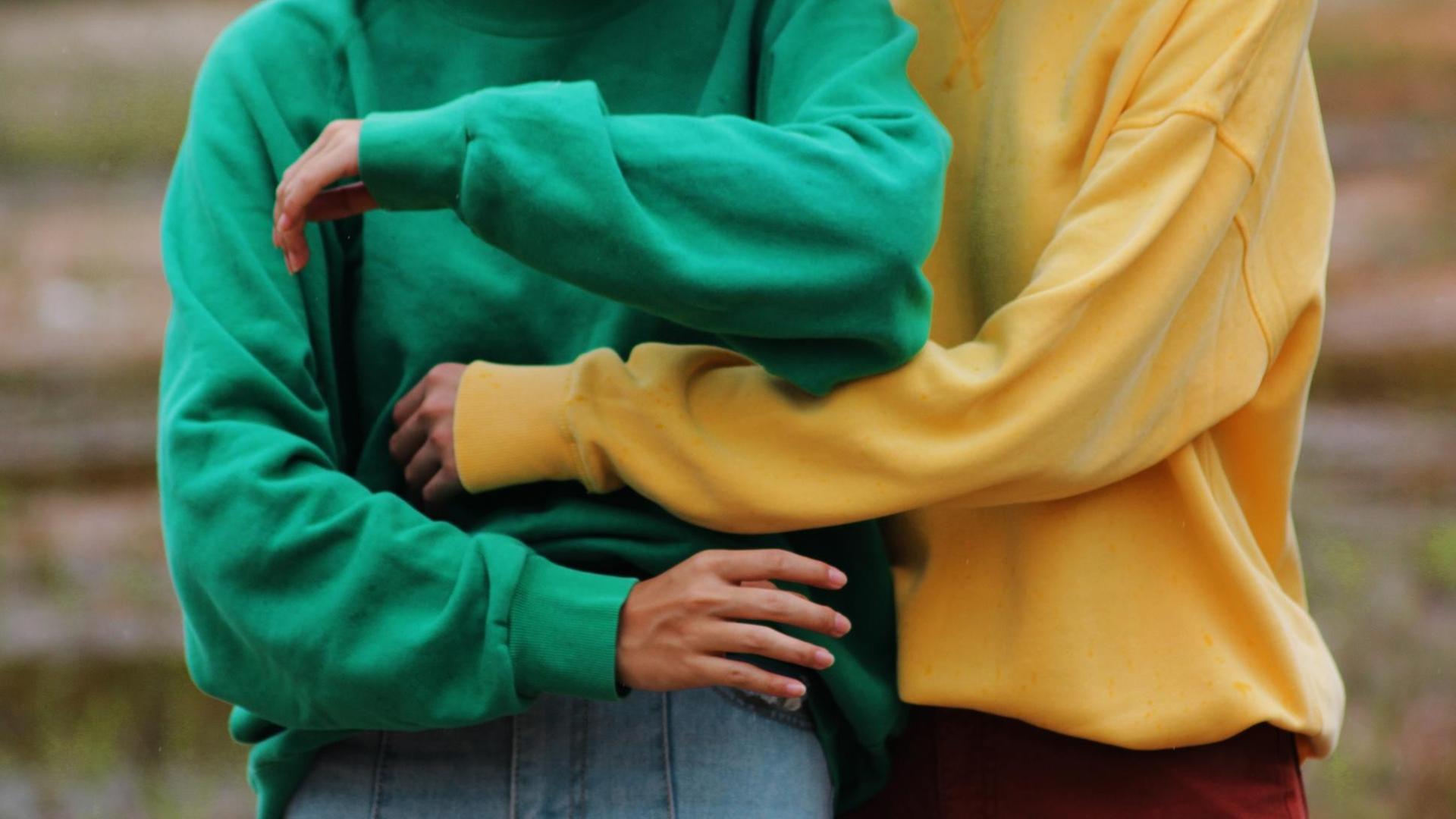 Detail von zwei Oberkörpern. Eine Person in gelbem Pullover umarmt eine Person in grünem Pullover. Detail von zwei Oberkörpern. Eine Person in gelbem Pullover umarmt eine Person in grünem Pullover.