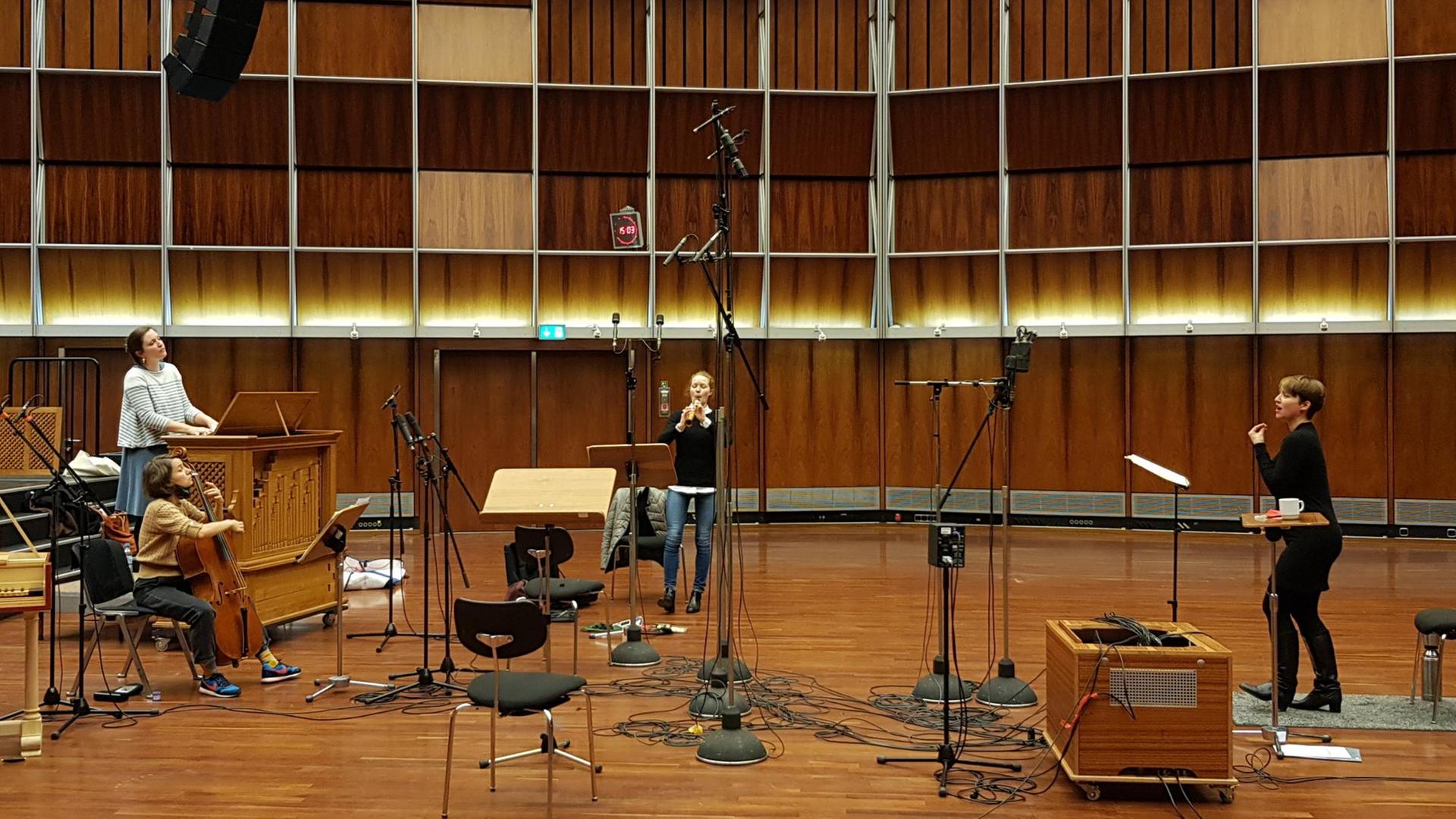 Im holzgetäfelten Deutschlandfunk Kammermusiksaal ist eine ganze Reihe von Standmikrophonen aufgebaut, die Sängerin steht rechts, sie trägt Stiefel und eine blaue Hose und einen blauen Pullover, links steht die Organistin an einer kleinen tragbaren Orgel, davor sitzt die Cellistin, in der Mitte des Bildes steht die Oboistin. Im holzgetäfelten Deutschlandfunk Kammermusiksaal ist eine ganze Reihe von Standmikrophonen aufgebaut, die Sängerin steht rechts, sie trägt Stiefel und eine blaue Hose und einen blauen Pullover, links steht die Organistin an einer kleinen tragbaren Orgel, davor sitzt die Cellistin, in der Mitte des Bildes steht die Oboistin.