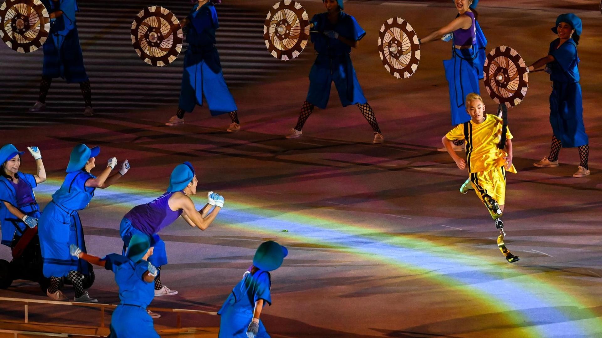 Ein Sportler mit einer Unterschenkelprothese läuft durch ein Spalier von jubelnden Statisten bei der Eröffnungsfeier der Paralympics 2020 Ein Sportler mit einer Unterschenkelprothese läuft durch ein Spalier von jubelnden Statisten bei der Eröffnungsfeier der Paralympics 2020