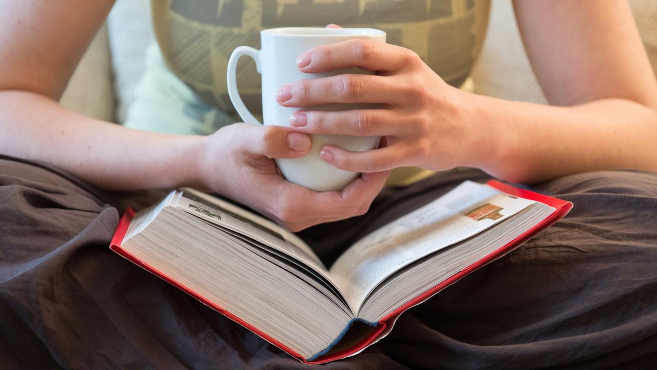 Hände einer Frau halten eine Tasse über einem dicken Buch.