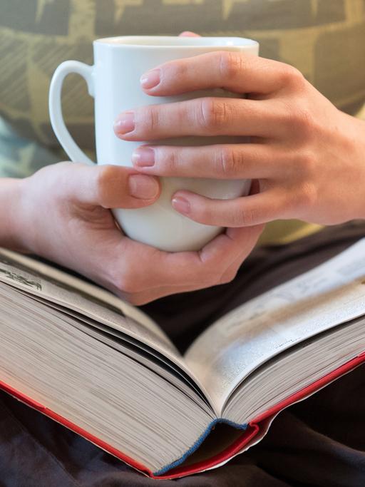 Hände einer Frau halten eine Tasse über einem dicken Buch.