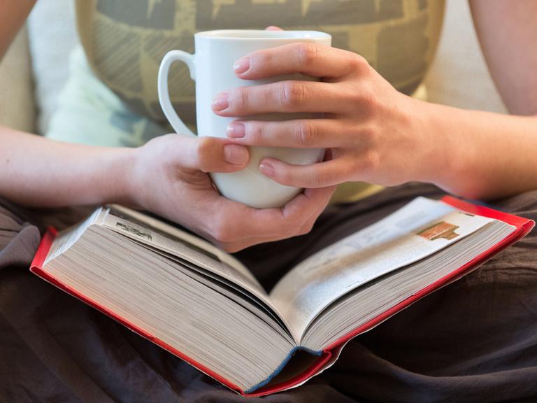 Hände einer Frau halten eine Tasse über einem dicken Buch.