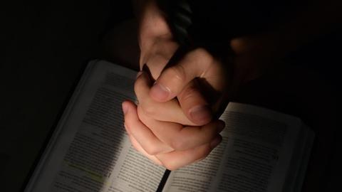 Detail von betenden Händen auf einer Bibel, auf die ein Lichtstahl fällt.
