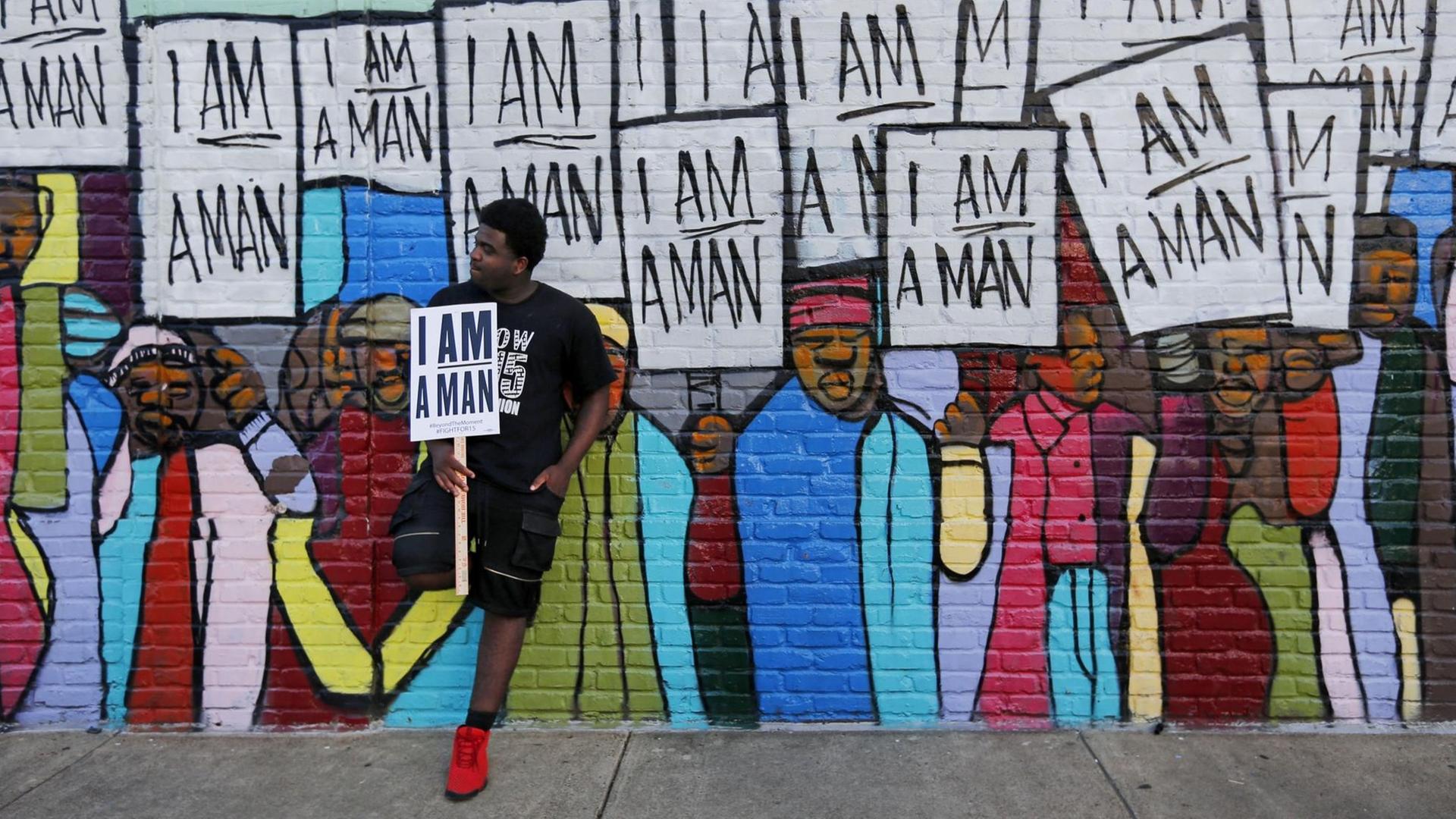 Ein Mann, mit afroamerikanischen Wurzeln, lehnt sich an ein Wandbild mit dem Schild "I am a Man" in der Hand. Ein Mann, mit afroamerikanischen Wurzeln, lehnt sich an ein Wandbild mit dem Schild "I am a Man" in der Hand.