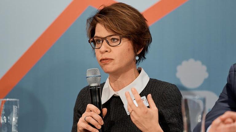 Eine junge Frau steht mit anderen Personen auf einem Podium und sagt etwas. Eine junge Frau steht mit anderen Personen auf einem Podium und sagt etwas.