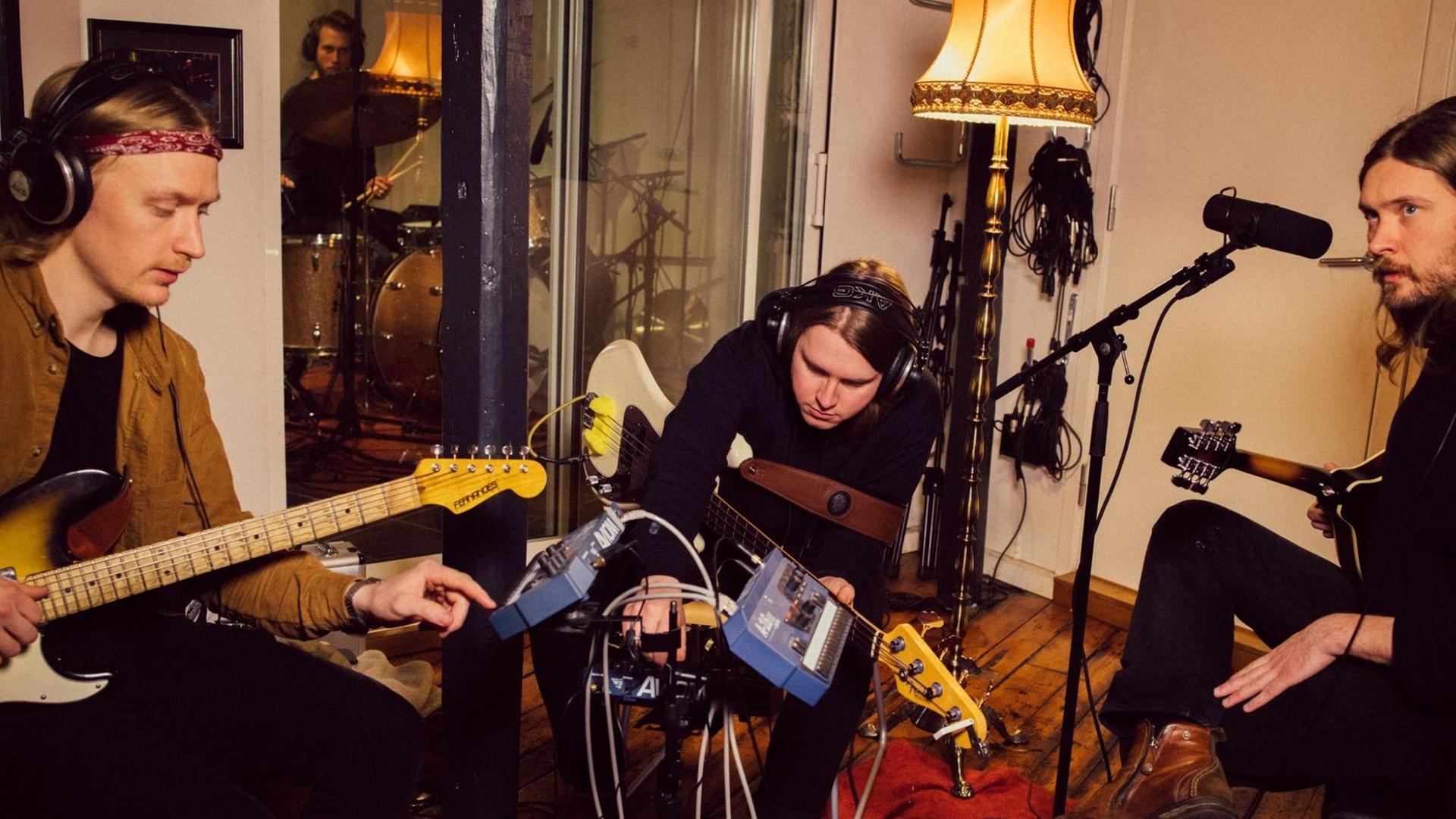Drei junge Männer sitzen mit E-Gitarren und E-Bass zusammen und tragen Kopfhörer, hinter einer Glasscheibe ein vierter Mann am Schlagzeug. Drei junge Männer sitzen mit E-Gitarren und E-Bass zusammen und tragen Kopfhörer, hinter einer Glasscheibe ein vierter Mann am Schlagzeug.