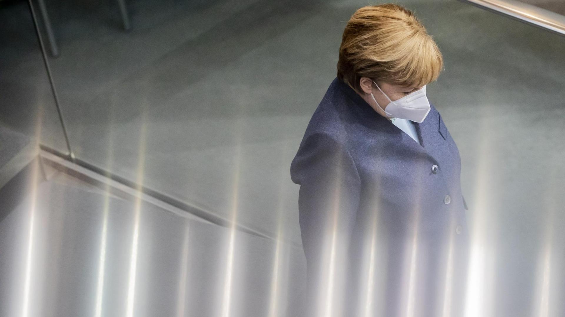 Angela Merkel trägt Maske und ist in einer Seitenansicht zu sehen. Vom unteren Rand treten irrisierende Lichteffeke ins Bild, die das Geschehen veruneindeutigen. Angela Merkel trägt Maske und ist in einer Seitenansicht zu sehen. Vom unteren Rand treten irrisierende Lichteffeke ins Bild, die das Geschehen veruneindeutigen.