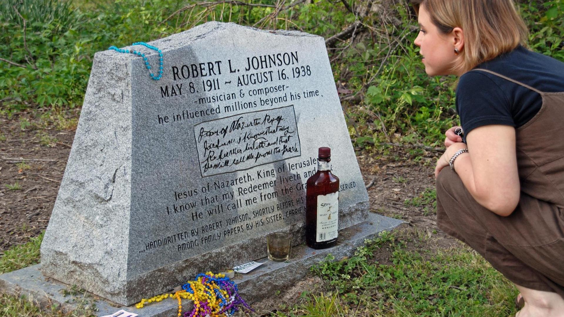 Ein Frau kniet vor dem Grabstein des Musikers Robert Johnson. Vor dem Grabstein steht eine braune Flasche und ein Glas. Daneben liegen bunte Ketten.