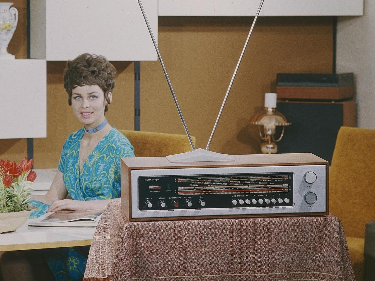 Eine junge Frau sitzt in einem Siebzigerjahre-Wohnzimmer an einem Tisch, im Vordergrund steht ein Radiogeraet REMA adagio. 