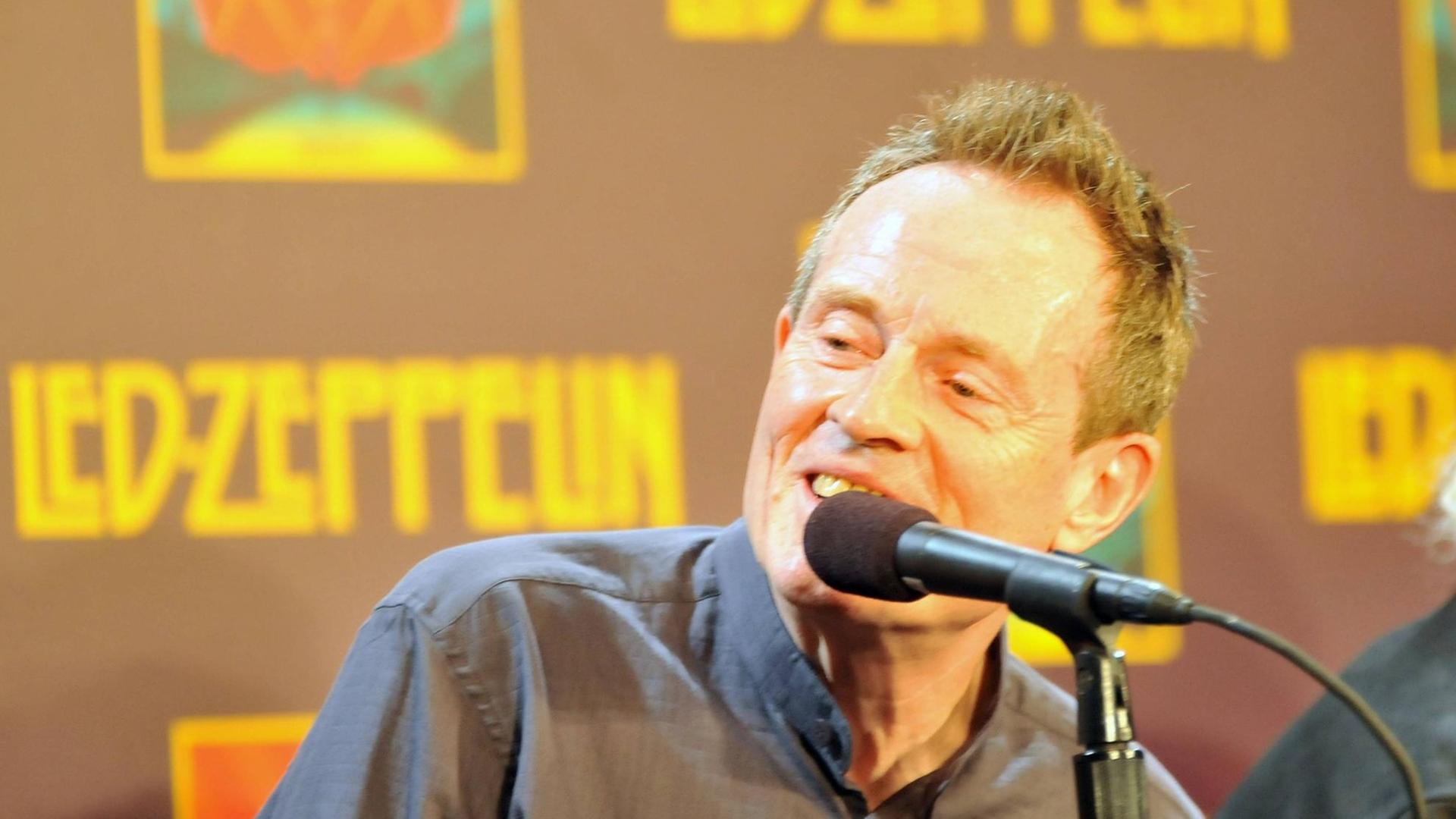Ein Mann mit kurzen Harren sitzt vor einem Mikrofon. Im Hintergrund befindet sich eine Plakatwand mit dem Schriftzug "Led Zeppelin".