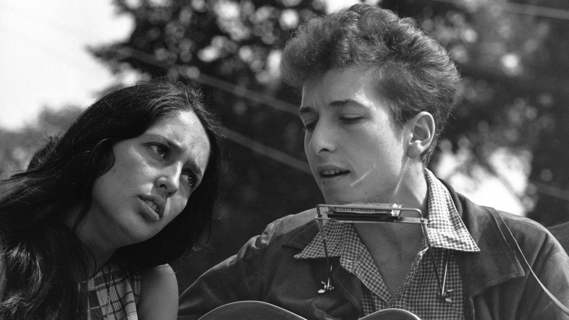 Ein Schwarzweiß-Foto zeigt eine Frau in Sommerkleid gelehnt an die Schulter eines Mannes mit Akustik-Gitarre und Mundharmonika Das Traumpaar des Folk: Joan Baez und Bob Dylan Hier 1963 am Rande des "Marschs auf Washington "1963