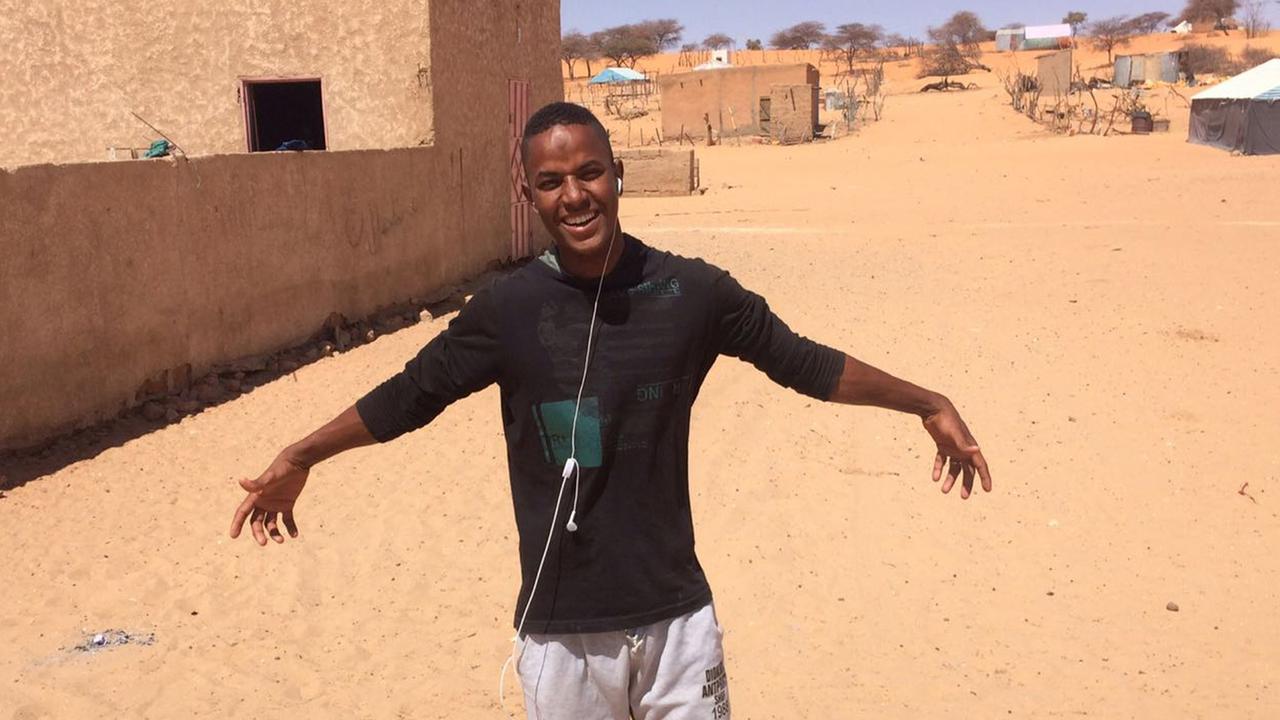 Ein junger Mann steht in einem Dorf auf einem gelben Sandboden, hat die Arme ausgebreitet und lächelt in die Kamera.