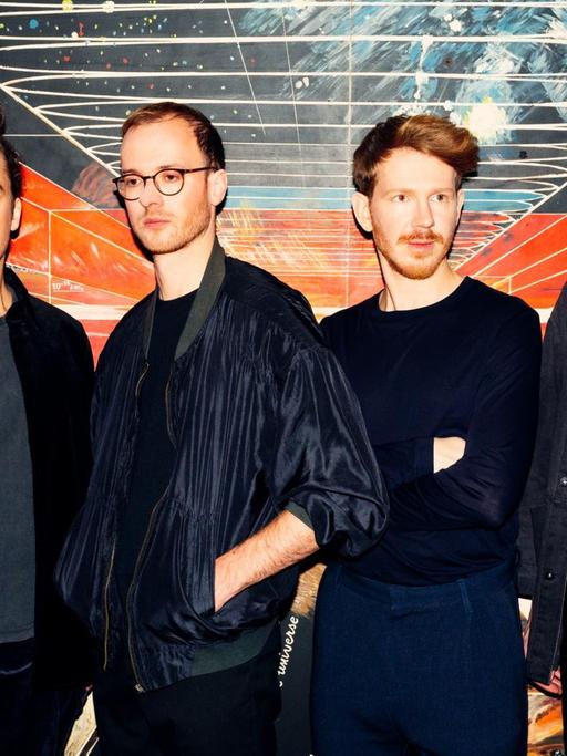 Vier schwarz gekleidete junge Musiker stehen vor einem bunten, surrealen Bild