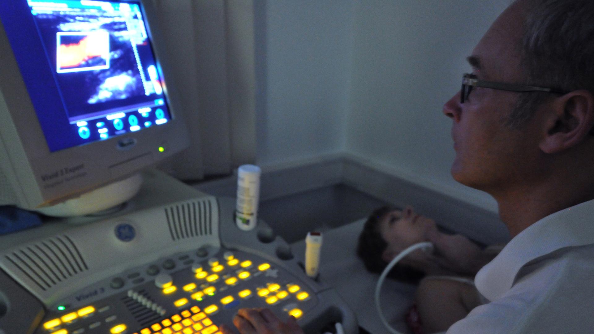 Ein Arzt sitzt in einem dunklen Raum vor einem Bildschirm eines Ultraschallgeräts, in der rechten Hand ein Ultraschallgerät, das an der Halsschlagader einer Patientin liegt. Ein Arzt sitzt in einem dunklen Raum vor einem Bildschirm eines Ultraschallgeräts, in der rechten Hand ein Ultraschallgerät, das an der Halsschlagader einer Patientin liegt.