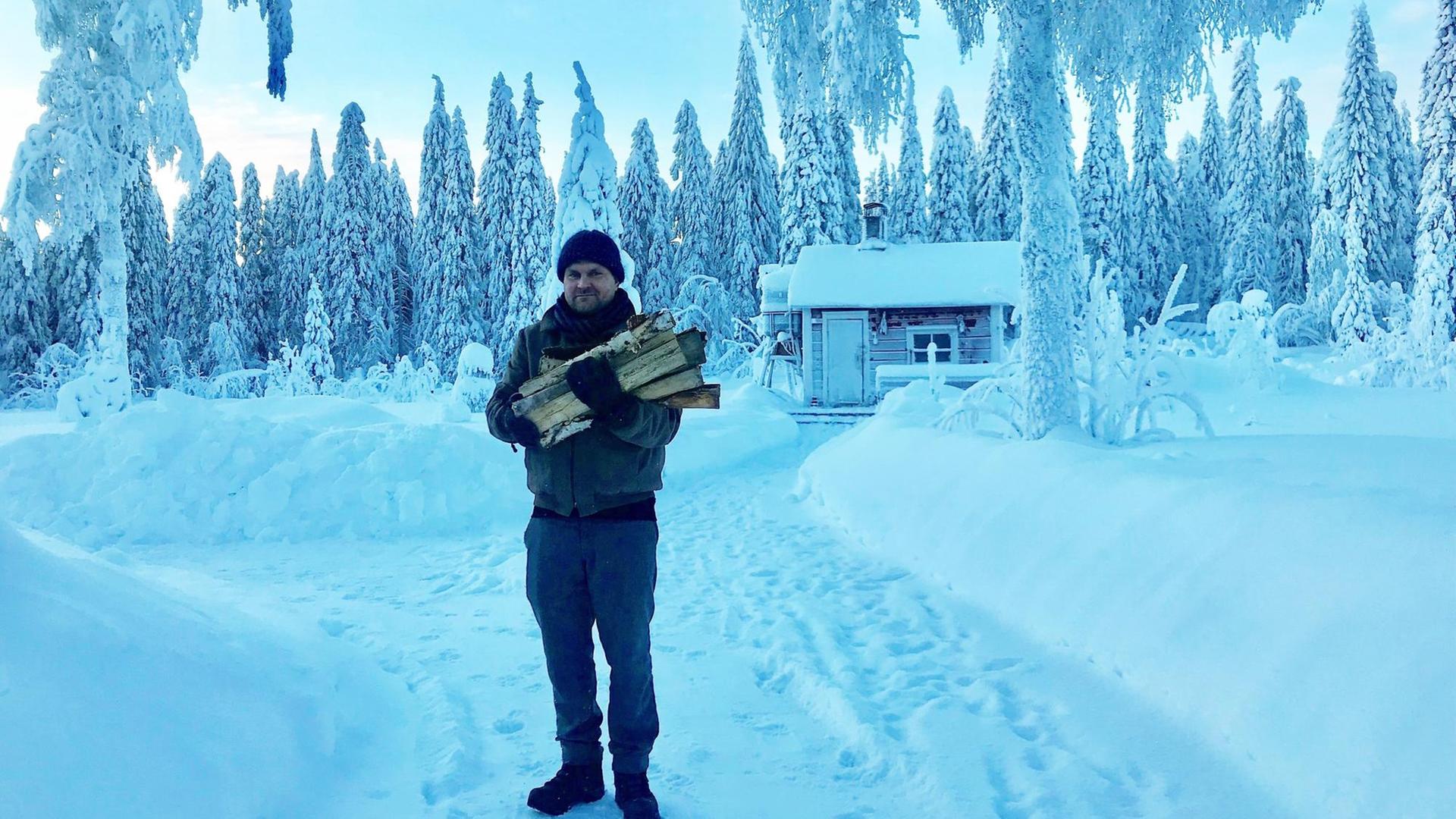 Der finnische Künstler Antti Majava steht mit Holzscheiten auf dem Arm in einer verschneiten Waldlandschaft vor einer Hütte