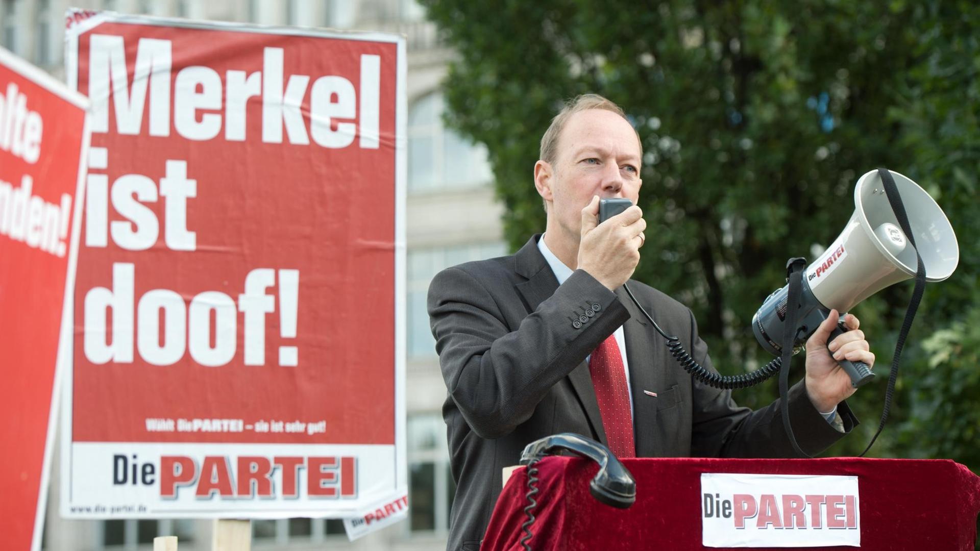 Martin Sonneborn spricht durch ein Megaphon, neben ihm ein Wahlplakat mit dem Slogan "Merkel ist doof!"