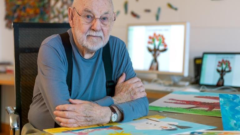 Kinderbuchautor Eric Carle sitzt mit Brille, Glatze und Bart an seinem Schreibtisch. Um ihn herum Zeichnungen.