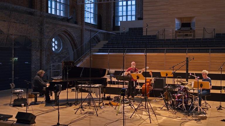 Drei Musiker sitzen mit großem Abstand auf einer Bühne in einem modernisierten Kirchenraum.