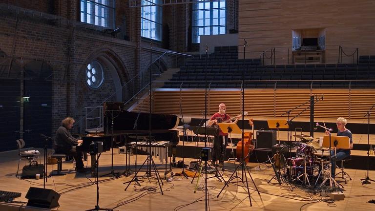 Drei Musiker sitzen mit großem Abstand auf einer Bühne in einem modernisierten Kirchenraum. Drei Musiker sitzen mit großem Abstand auf einer Bühne in einem modernisierten Kirchenraum.