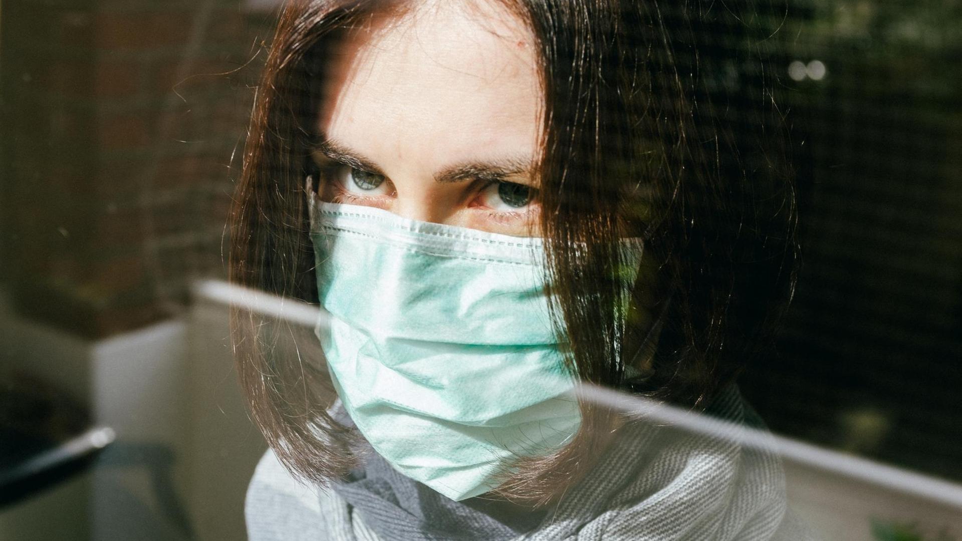 Großaufnahme einer Frau mit Schutzmaske, die aus einem Fenster schaut. Großaufnahme einer Frau mit Schutzmaske, die aus einem Fenster schaut.