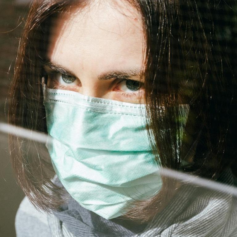 Großaufnahme einer Frau mit Schutzmaske, die aus einem Fenster schaut.