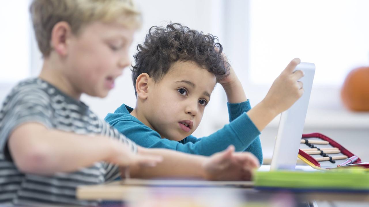 Zwei Kinder sitzen an einer Schulbank und arbeiten jeweils mit einem Tablet. Zwei Kinder sitzen an einer Schulbank und arbeiten jeweils mit einem Tablet.