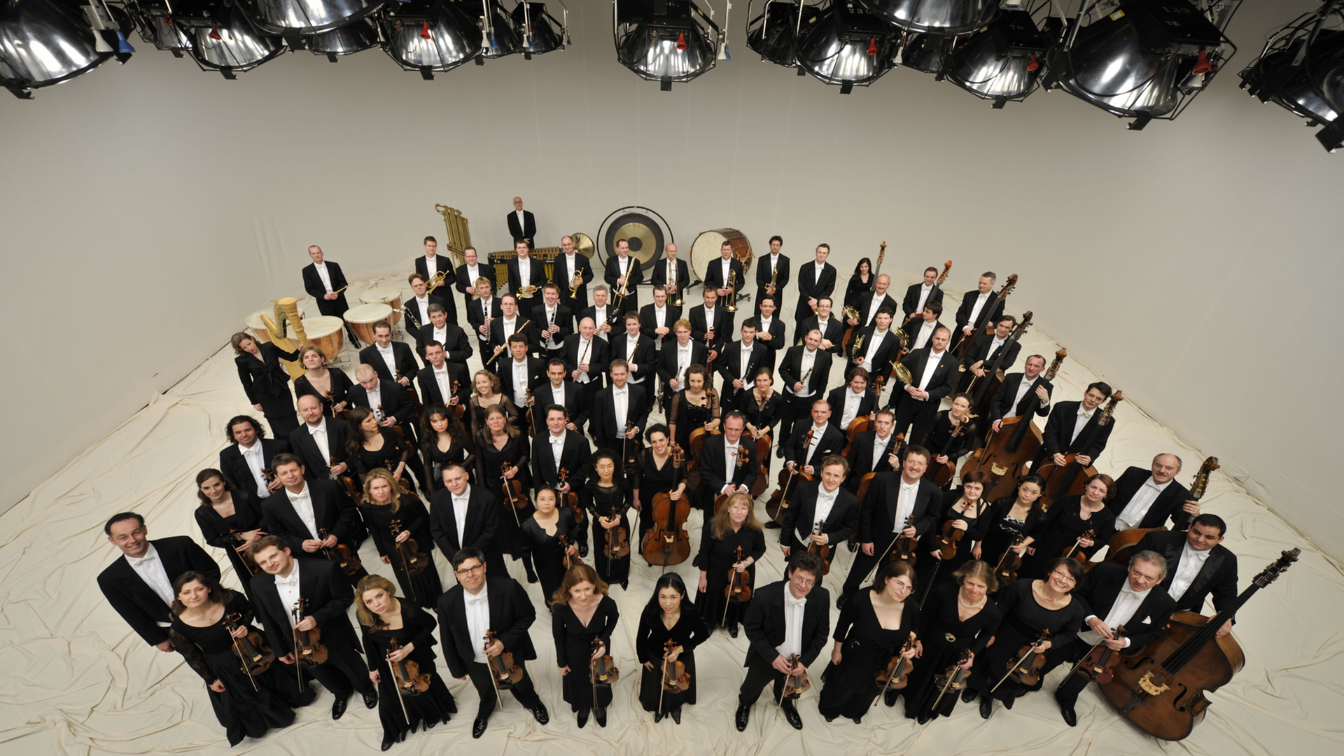 Die Mitglieder des Orchesters stehen mit ihren Instrumenten in einem großen Raum und schauen nach oben in die Kamera. Die Mitglieder des Orchesters stehen mit ihren Instrumenten in einem großen Raum und schauen nach oben in die Kamera.