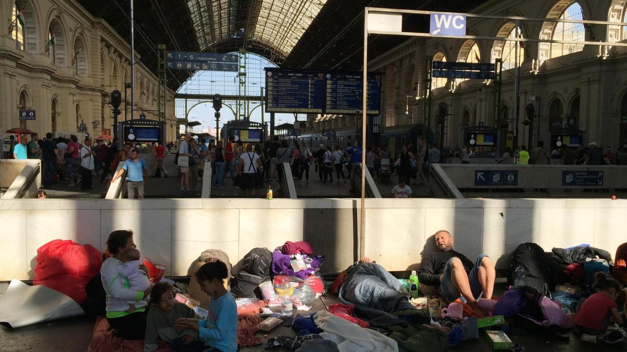 Flüchtlinge sitzen auf Matten und an eine Mauer gelehnt in einer Bahnhofshalle.