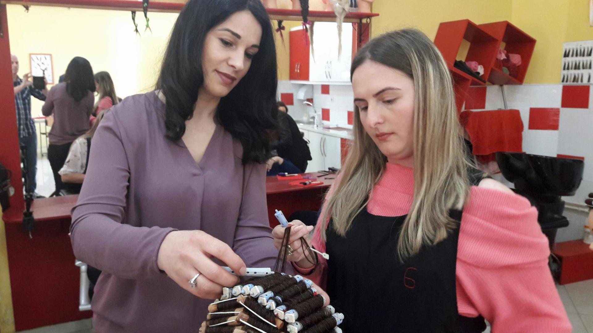 Zwei Frauen drehen Lockenwickler an einem Übungskopf mit Perücke ein Zwei Frauen drehen Lockenwickler an einem Übungskopf mit Perücke ein
