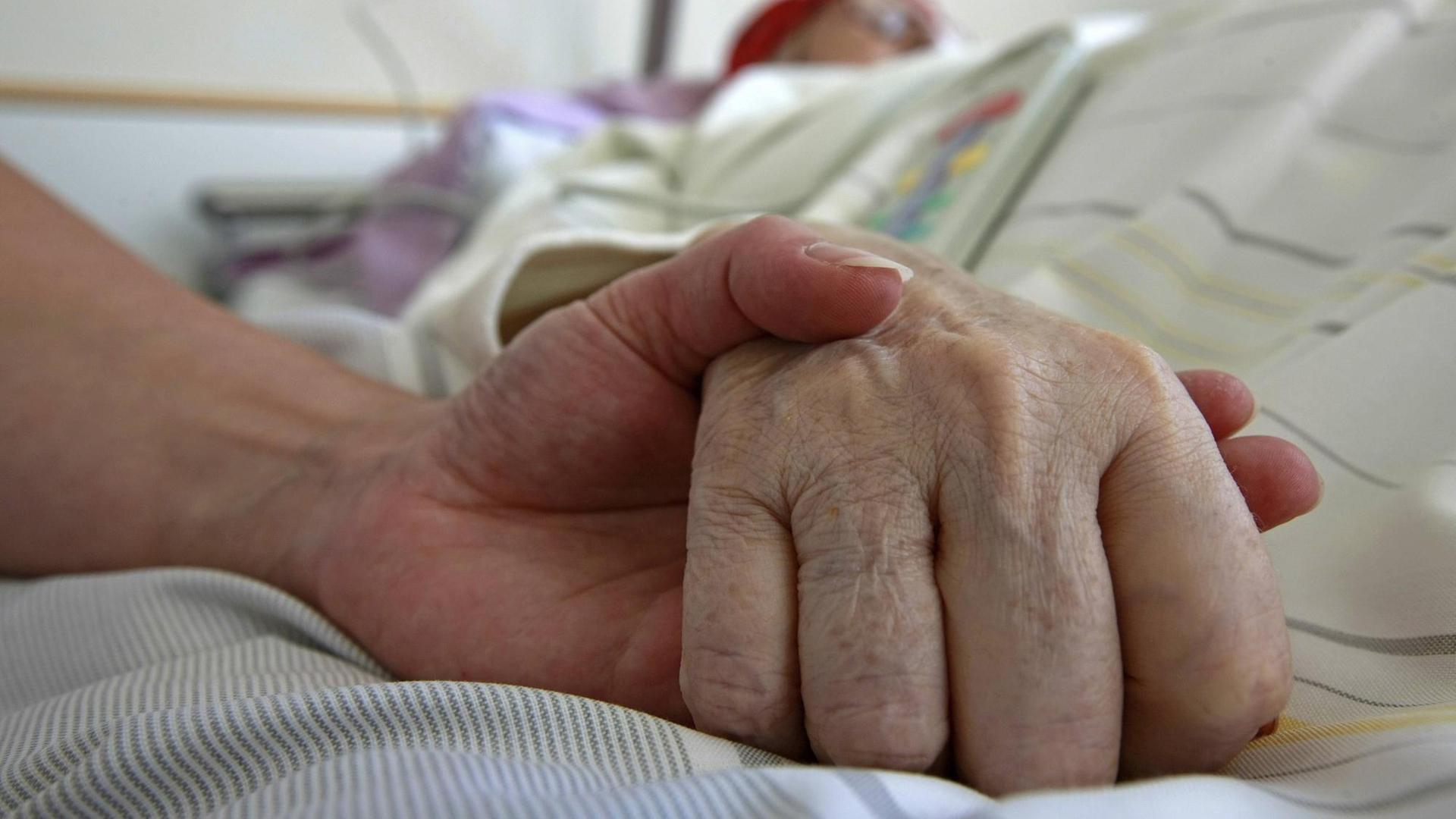 Großaufnahme einer Hand, die auf einem Krankenbett die Hand eines Intensivpatienten hält. Großaufnahme einer Hand, die auf einem Krankenbett die Hand eines Intensivpatienten hält.