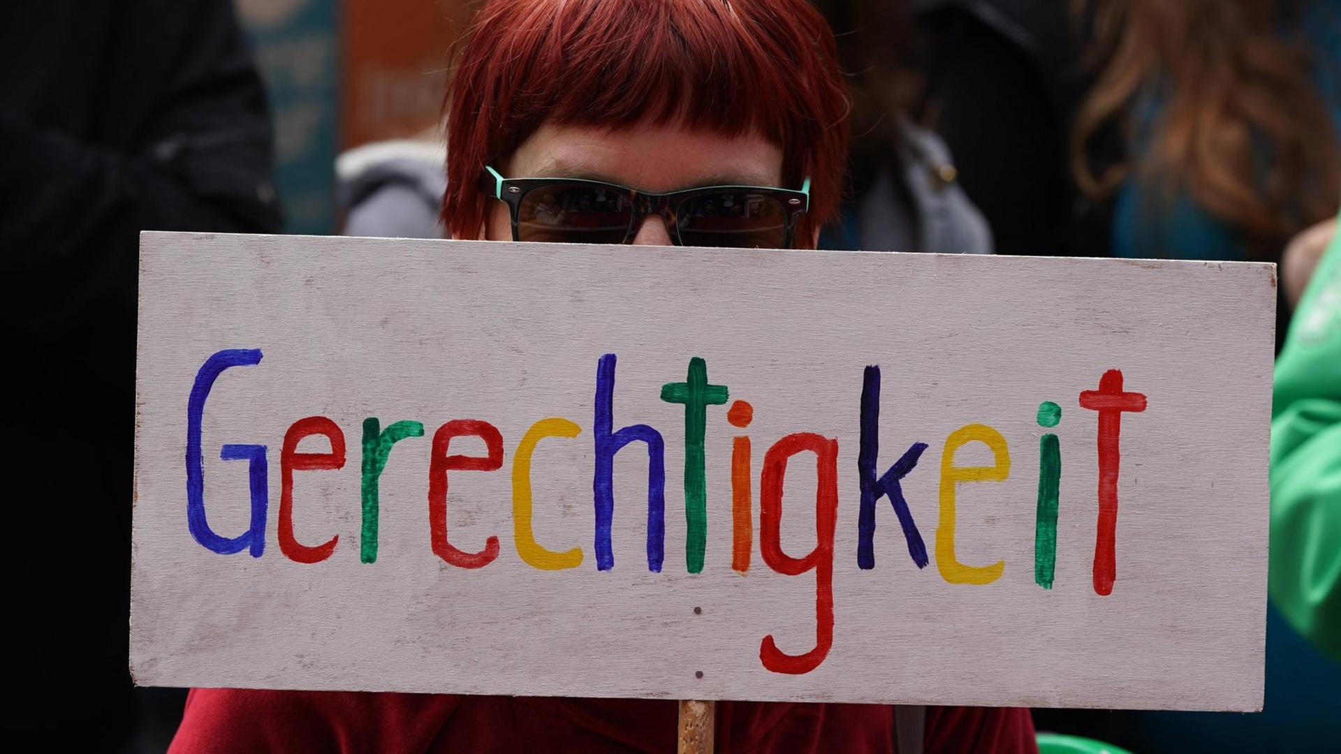 Das Bild zeigt eine Frau, die ein Schild mit der Aufschrift Gerechtigkeit vor ihr Gesicht hält. Das Bild zeigt eine Frau, die ein Schild mit der Aufschrift Gerechtigkeit vor ihr Gesicht hält.
