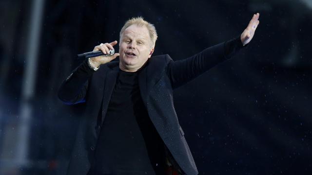 Herbert Grönemeyer singend und mit ausgestrecktem Arm auf der Bühne, dunkle Kleidung, dunkler Hintergrund, 2015