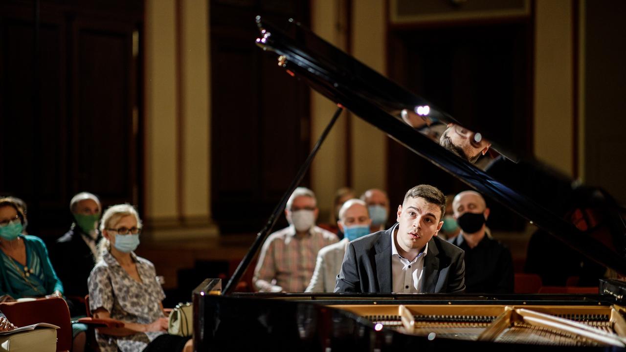 Der Pianist wurde von der hinteren Seite des schwarzen Flügels aufgenommen. Der Deckel ist aufgestellt, der junge Mann trägt ein weißes Hemd und ein graue Jacket, er hat kurze braune Haare, er schaut nach oben. Der Pianist wurde von der hinteren Seite des schwarzen Flügels aufgenommen. Der Deckel ist aufgestellt, der junge Mann trägt ein weißes Hemd und ein graue Jacket, er hat kurze braune Haare, er schaut nach oben.