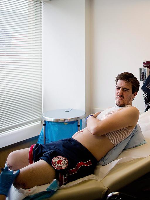 Jeff Bauman liegt nur mit kurzer Hose und Shirt bekleidet auf einer Liege in einer Reha-Klinik, ihm fehlen beide Unterschenkel, sein Gesicht zeigt Erschöpfung.
