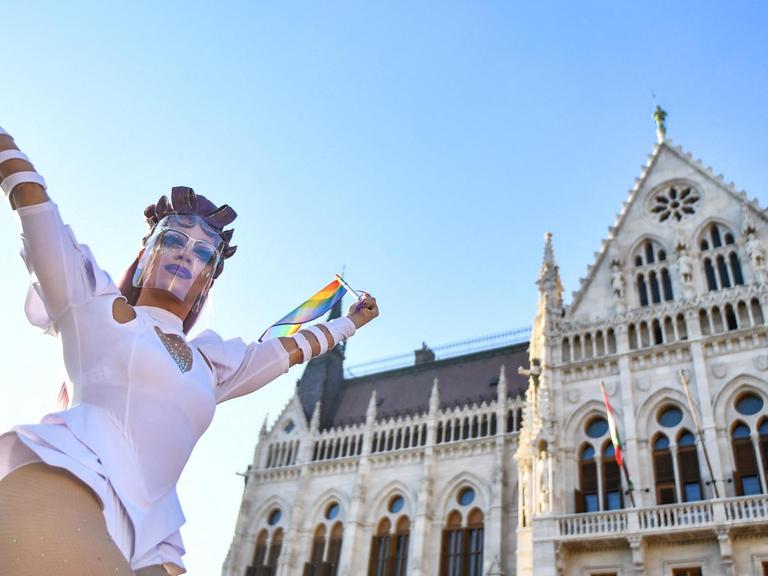 Eine Person mit sehr langen Fingernägeln und einem eng anliegenden weissen Anzug posiert mit einer Regenbogenfahne vor dem Parlament.