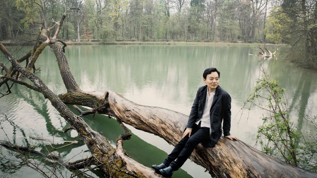 William Youn sitzt auf einem Baumstamm, der quer in einen See ragt William Youn sitzt auf einem Baumstamm, der quer in einen See ragt