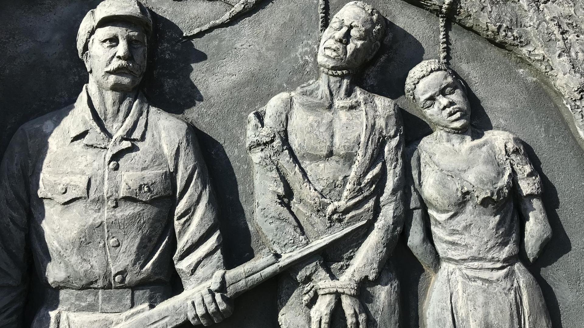Denkmal in Windhoek für den von deutschen Kolonialtruppen begangenen Völkermord an den Herero und Nama: An einem Ast hängen die an den Händen gefesselten Leichen eines Mannes und einer Frau. Daneben steht ein kräftiger Mann mit einem Gewehr. Denkmal in Windhoek für den von deutschen Kolonialtruppen begangenen Völkermord an den Herero und Nama: An einem Ast hängen die an den Händen gefesselten Leichen eines Mannes und einer Frau. Daneben steht ein kräftiger Mann mit einem Gewehr.