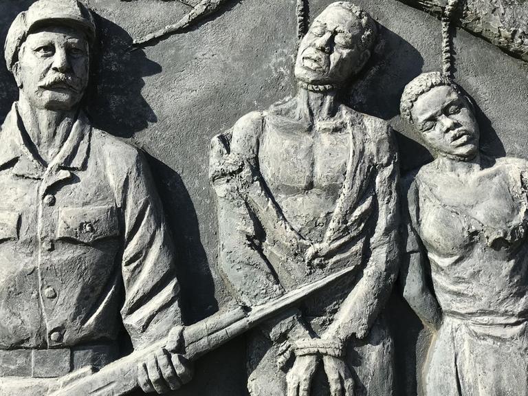 Denkmal in Windhoek für den von deutschen Kolonialtruppen begangenen Völkermord an den Herero und Nama: An einem Ast hängen die an den Händen gefesselten Leichen eines Mannes und einer Frau. Daneben steht ein kräftiger Mann mit einem Gewehr.