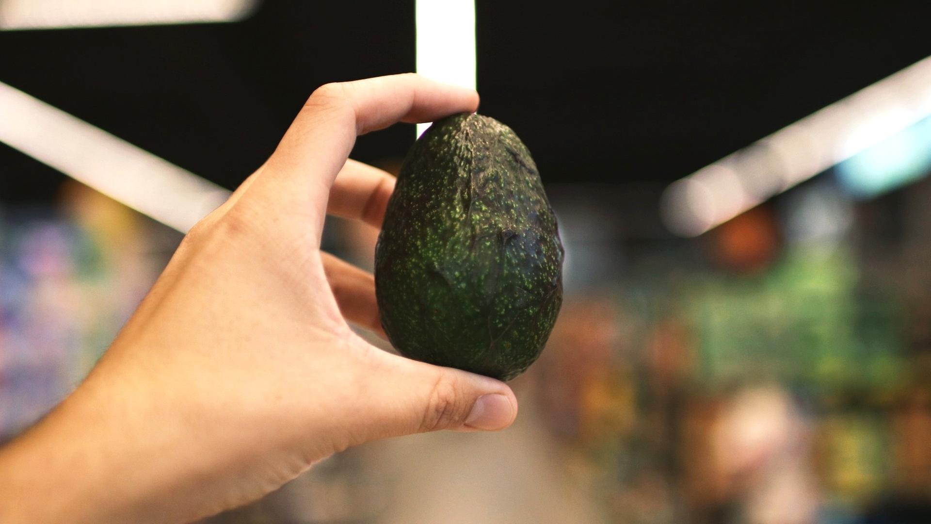 Eine Person hält eine Avocado. Im Hintergrund, in der Unschärfe ein Supermarkt. Eine Person hält eine Avocado. Im Hintergrund, in der Unschärfe ein Supermarkt.