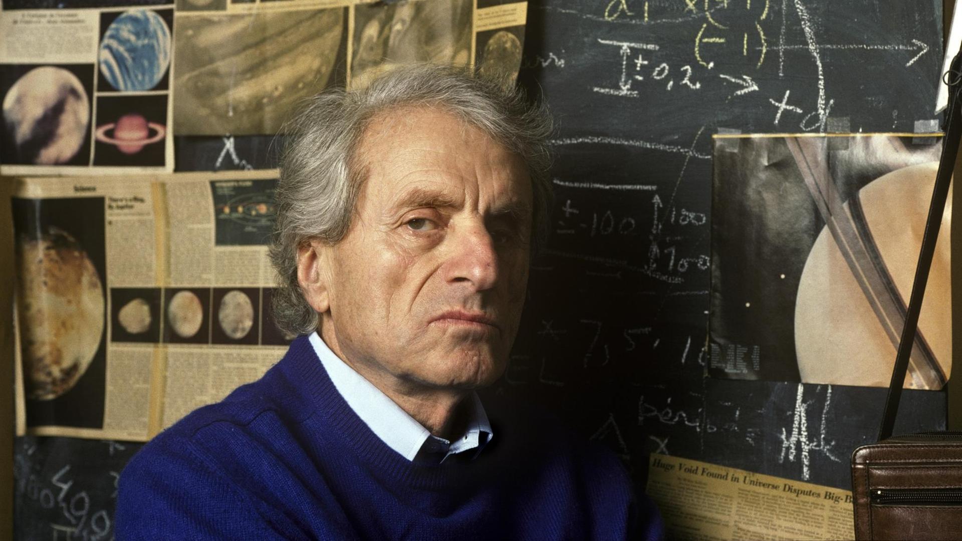 Iannis Xenakis steht an einer Tafel auf der Zeitungsausschnitte mit Planeten zu sehen sind. Iannis Xenakis steht an einer Tafel auf der Zeitungsausschnitte mit Planeten zu sehen sind.