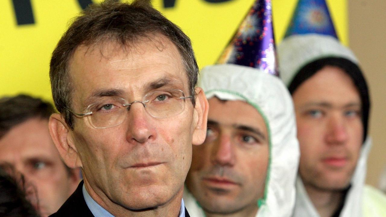 Piebalgs blickt genervt. Hinter ihm Demonstranten in weißen Schutzanzügen vor einem gelben Transparent, auf dem man das Wort "Power!" lesen kann. Piebalgs blickt genervt. Hinter ihm Demonstranten in weißen Schutzanzügen vor einem gelben Transparent, auf dem man das Wort "Power!" lesen kann.