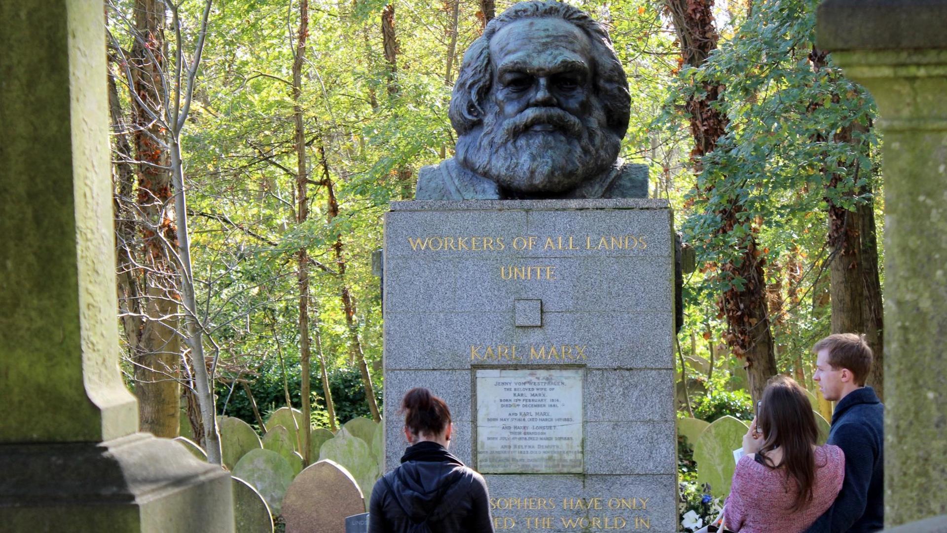 Das Bild zeigt den massiven Grabstein mit der Marmorplatte und der Marx-Büste darauf von vorne. Dahinter Bäume, vor dem Grab drei Menschen. Das Bild zeigt den massiven Grabstein mit der Marmorplatte und der Marx-Büste darauf von vorne. Dahinter Bäume, vor dem Grab drei Menschen.