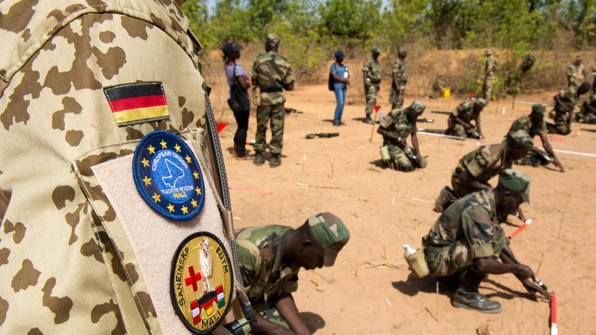 Die Schulter eines Bundeswehrsoldaten in Uniform ist zu sehen, dahinter knien Pioniere der Armee Malis. Die Schulter eines Bundeswehrsoldaten in Uniform ist zu sehen, dahinter knien Pioniere der Armee Malis.