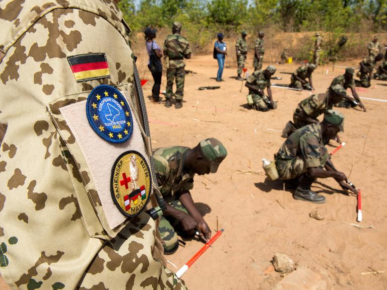 Die Schulter eines Bundeswehrsoldaten in Uniform ist zu sehen, dahinter knien Pioniere der Armee Malis.