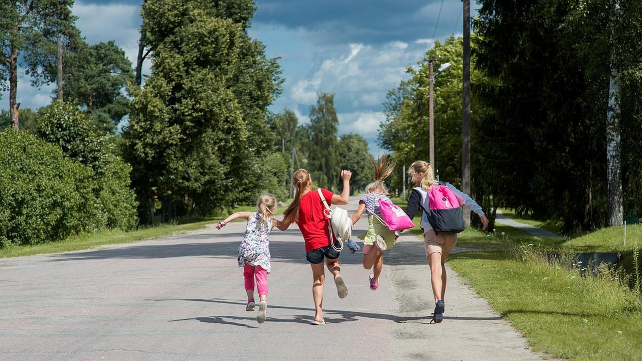 Vier Mädchen springen Hand in Hand auf einer Straße. Sie sind von hinten zu sehen.