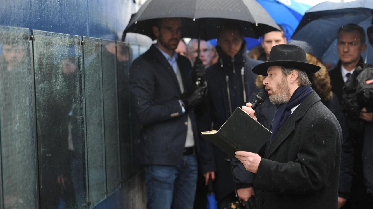 An der Tausend-Freunde-Mauer vor der Veltins-Arena in Gelsenkirchen betet Rabbiner Chaim Kornblum stehend vor der Gedenktafel. Im Hintergrund Spieler und Fans des FC Schalke 04. An der Tausend-Freunde-Mauer vor der Veltins-Arena in Gelsenkirchen betet Rabbiner Chaim Kornblum stehend vor der Gedenktafel. Im Hintergrund Spieler und Fans des FC Schalke 04.