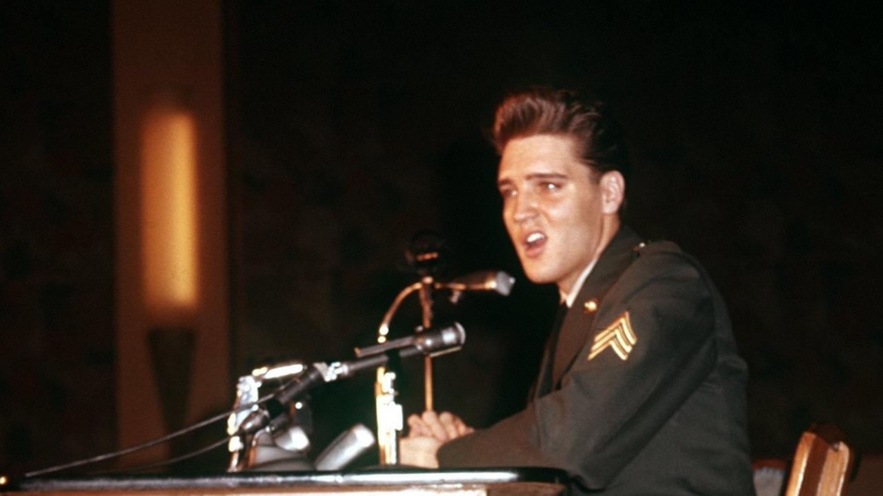Elvis Presley sitzt an einem Tisch, auf dem Mikrofone stehen.