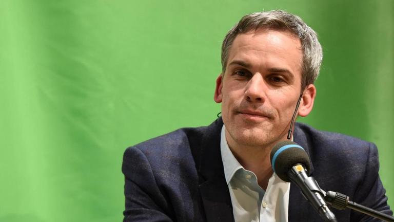 Stephan Thome sitzt an einem Tisch vor grünem Hintergrund, vor ihm ein Mikro. Er trägt ein dunkles Sakko und ein helles Hemd, ohne Krawatte. Stephan Thome sitzt an einem Tisch vor grünem Hintergrund, vor ihm ein Mikro. Er trägt ein dunkles Sakko und ein helles Hemd, ohne Krawatte.