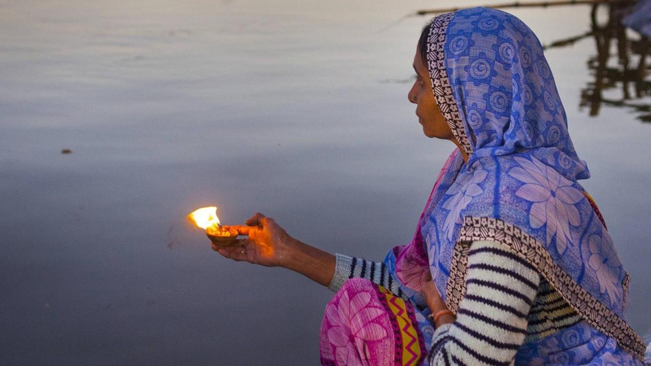 Eine Frau sitzt während des hinduistischen Festes Maha Kumbh Mela am Flussufer und hält eine brennende Gabe in der Hand.