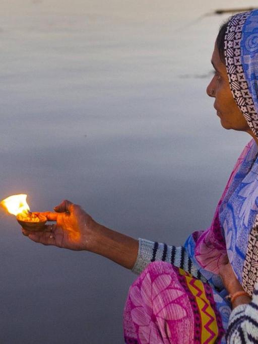 Eine Frau sitzt während des hinduistischen Festes Maha Kumbh Mela am Flussufer und hält eine brennende Gabe in der Hand.